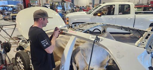 A person in a garage works on a vehicle with the hood open. A white truck is in the background.