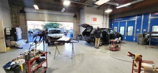 Inside a car body shop, a car is being worked on with open doors and a person standing near it.