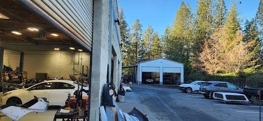 A car repair shop with open garage doors. Cars and equipment are visible outside and inside the shop. Trees and blue sky in the background.