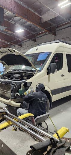 A person works on a beige van in a garage. The hood is open, and tools are nearby.