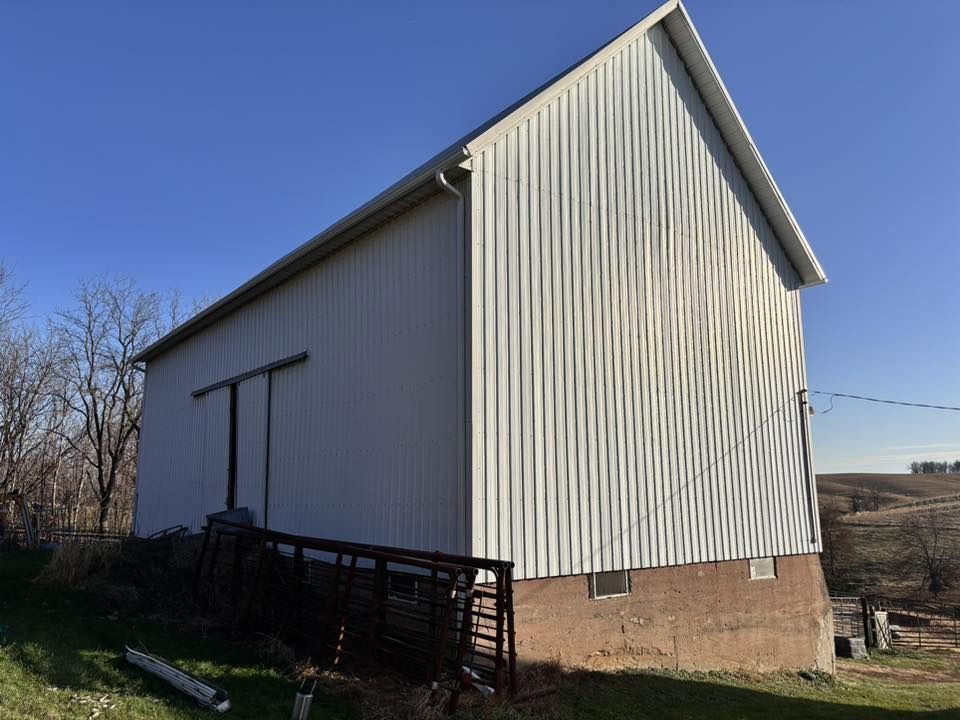 White metal-sided barn with a dark sliding door, set on a stone foundation against a clear blue sky.