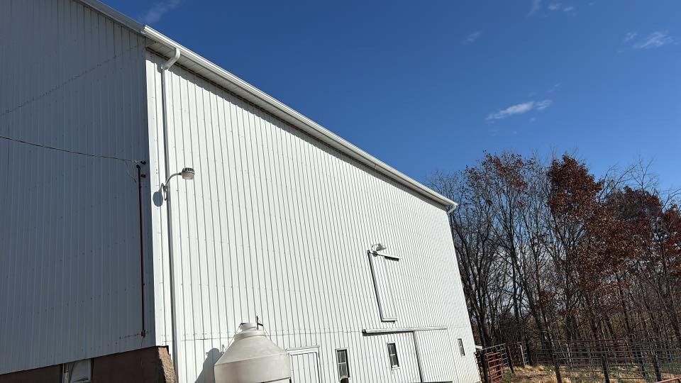 A white metal building with a gutter system, exterior light, and a small door against a clear blue sky and treeline.
