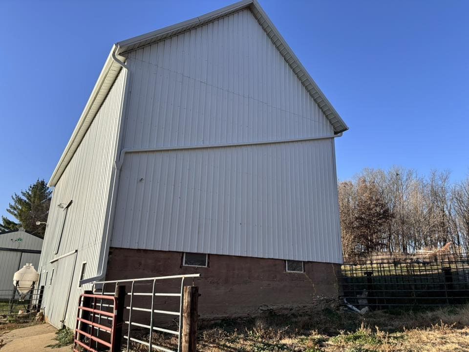 A large barn with white vertical metal siding over a brown foundation under a clear blue sky, next to a metal gate.