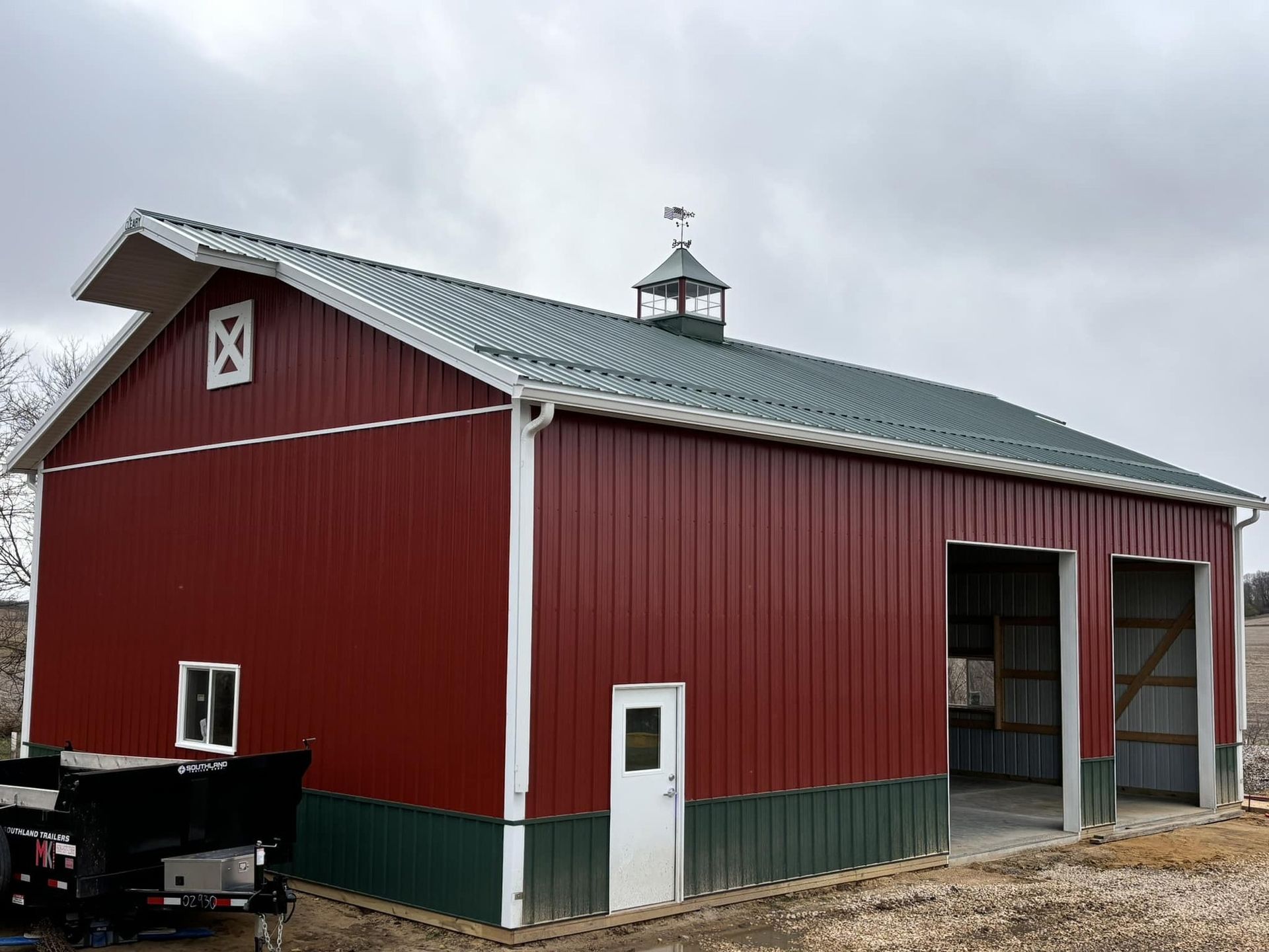 A red barn with green trim, a metal roof, a cupola, and two open bays, set against a cloudy sky.
