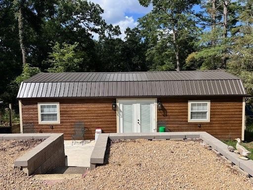 A brown wooden shed with a metal roof and double doors, situated in a yard with gravel and low stone walls.