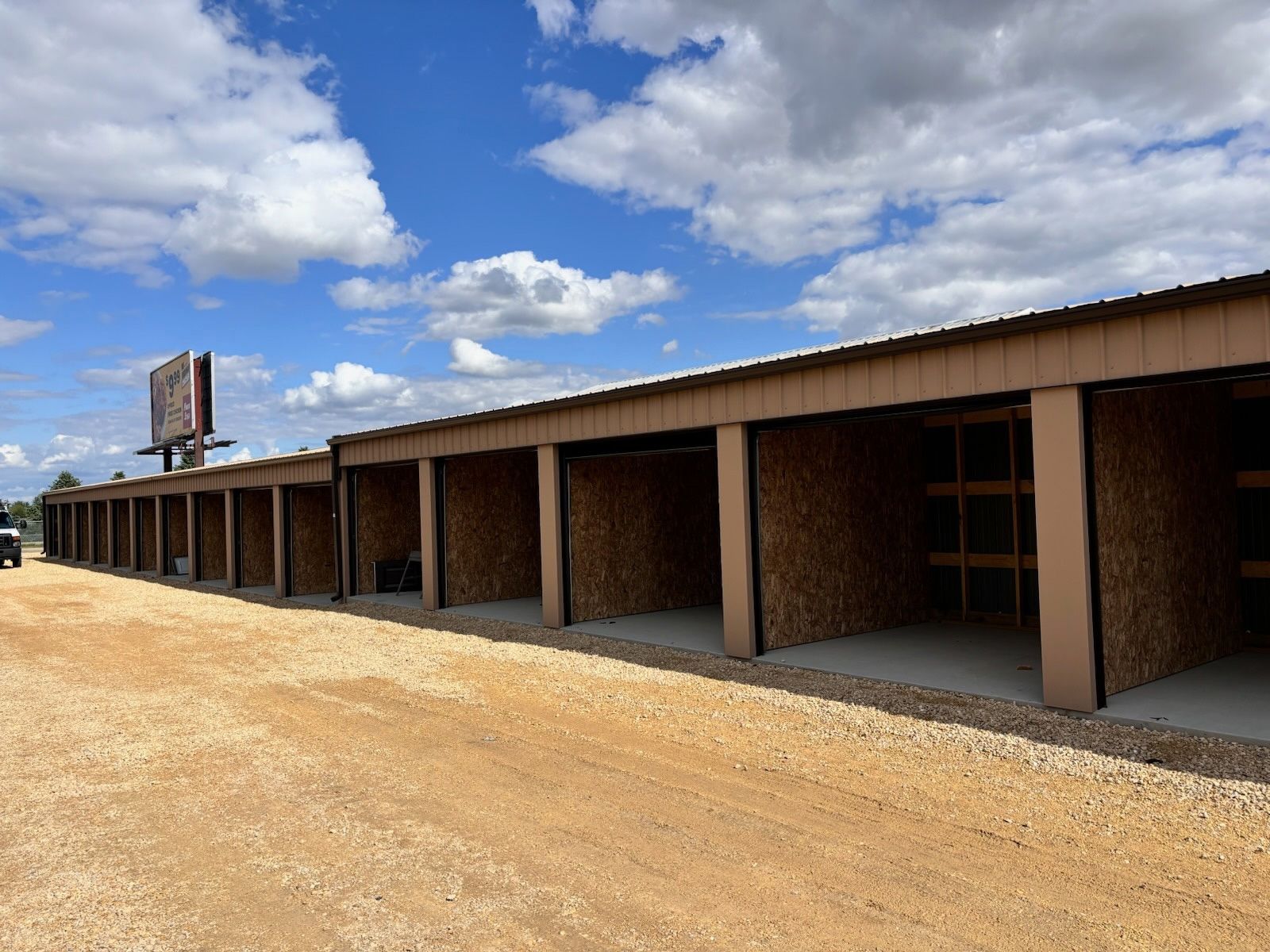 A long row of empty outdoor self-storage units with wooden walls and concrete floors under a bright, cloudy blue sky.
