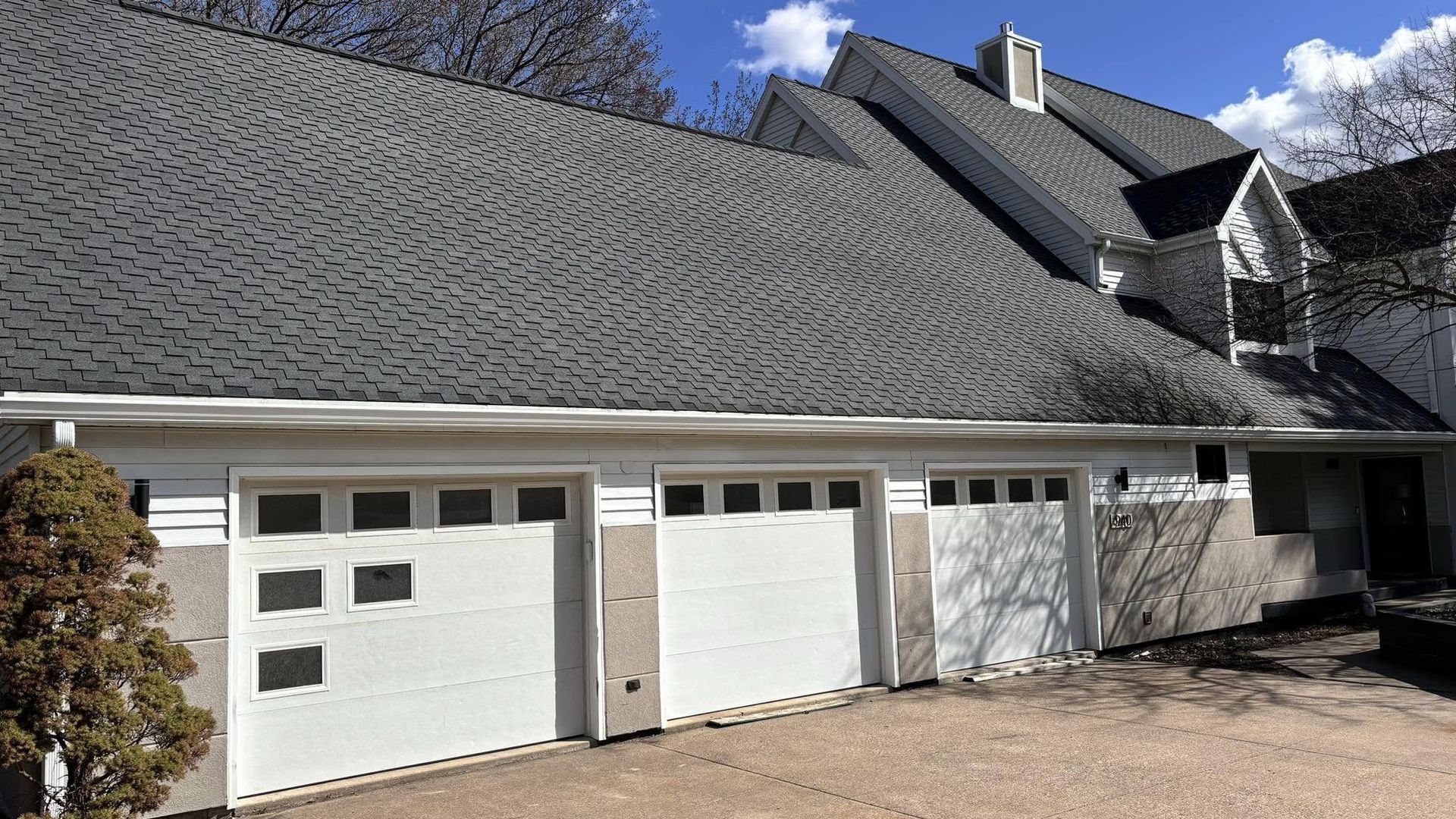 Three white garage doors on a house with a grey, shingled roof under a bright blue sky.