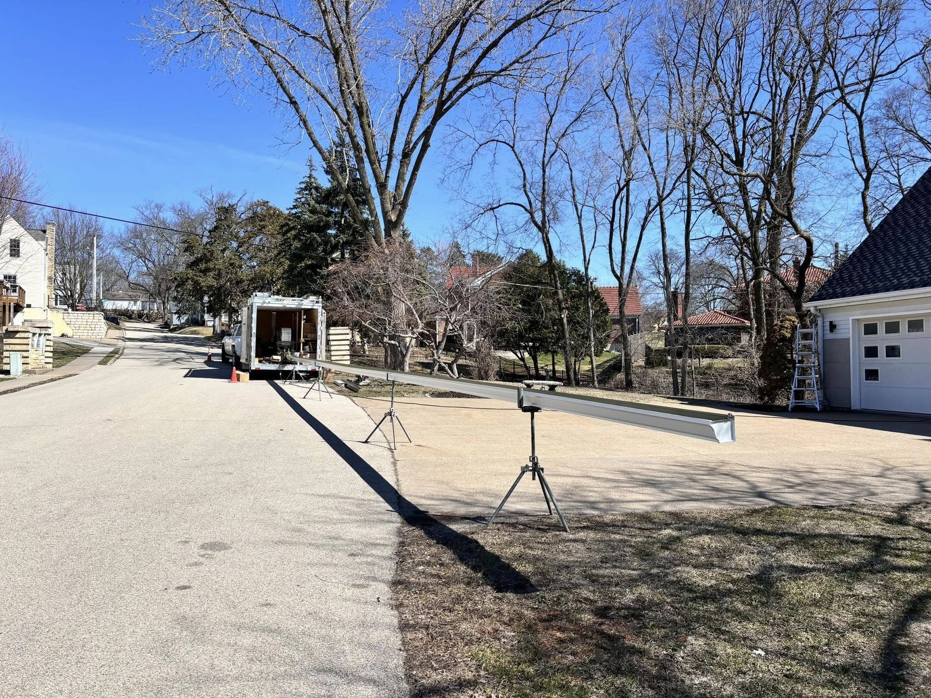 A long grey steel beam supported by tripods sits on a residential driveway near a parked trailer on a sunny day.