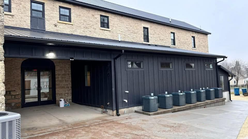 A stone building with a dark metal-clad ground floor, an entrance, and a row of outdoor HVAC units along the side.