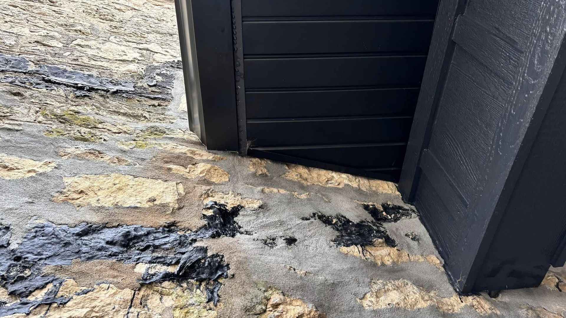 A close-up view of black wood siding meeting a textured, light-colored stone wall with black residue on the ground.