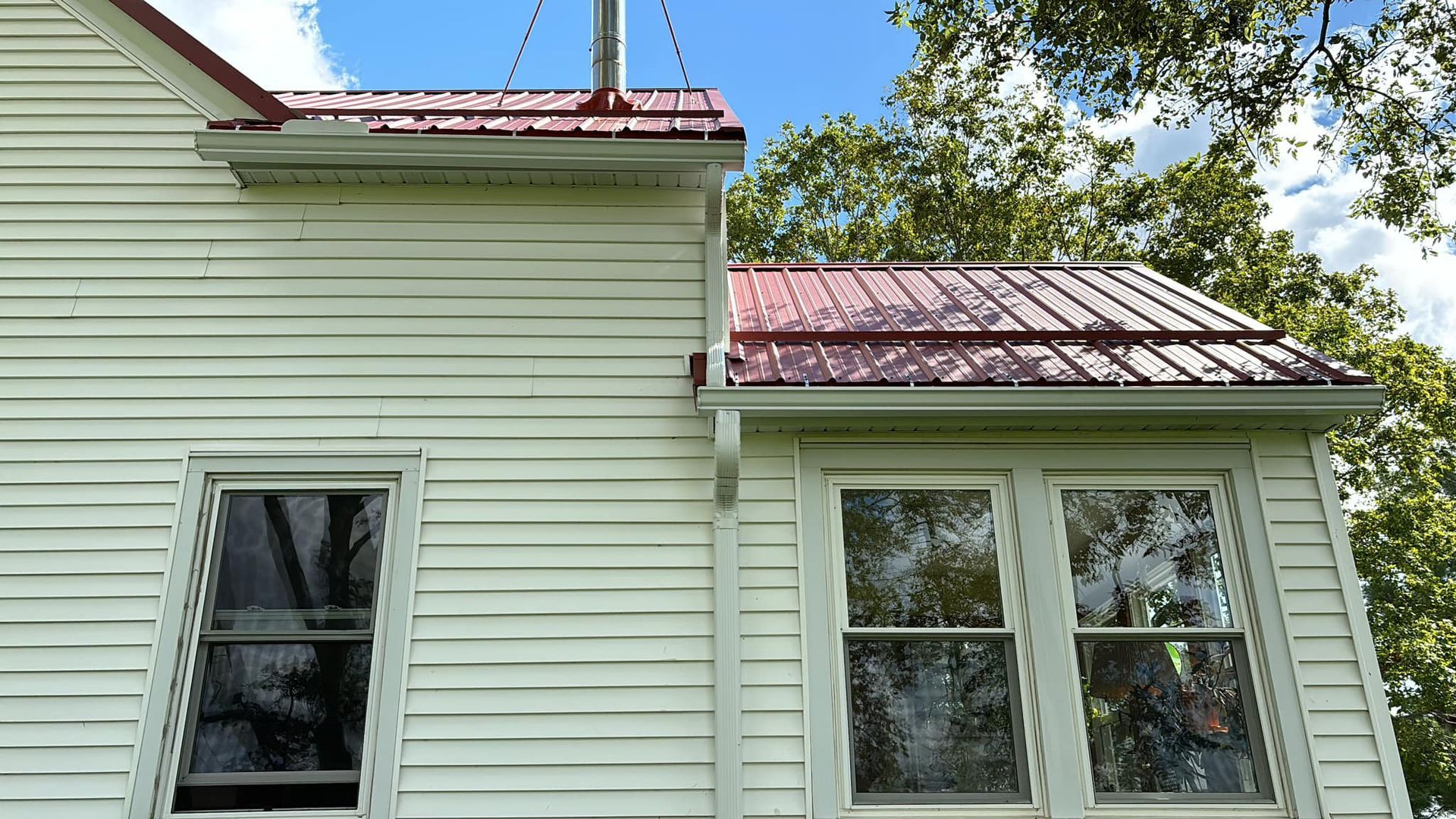 Exterior view of a house with pale yellow siding, two windows, and a red metal roof under a blue sky with green trees.