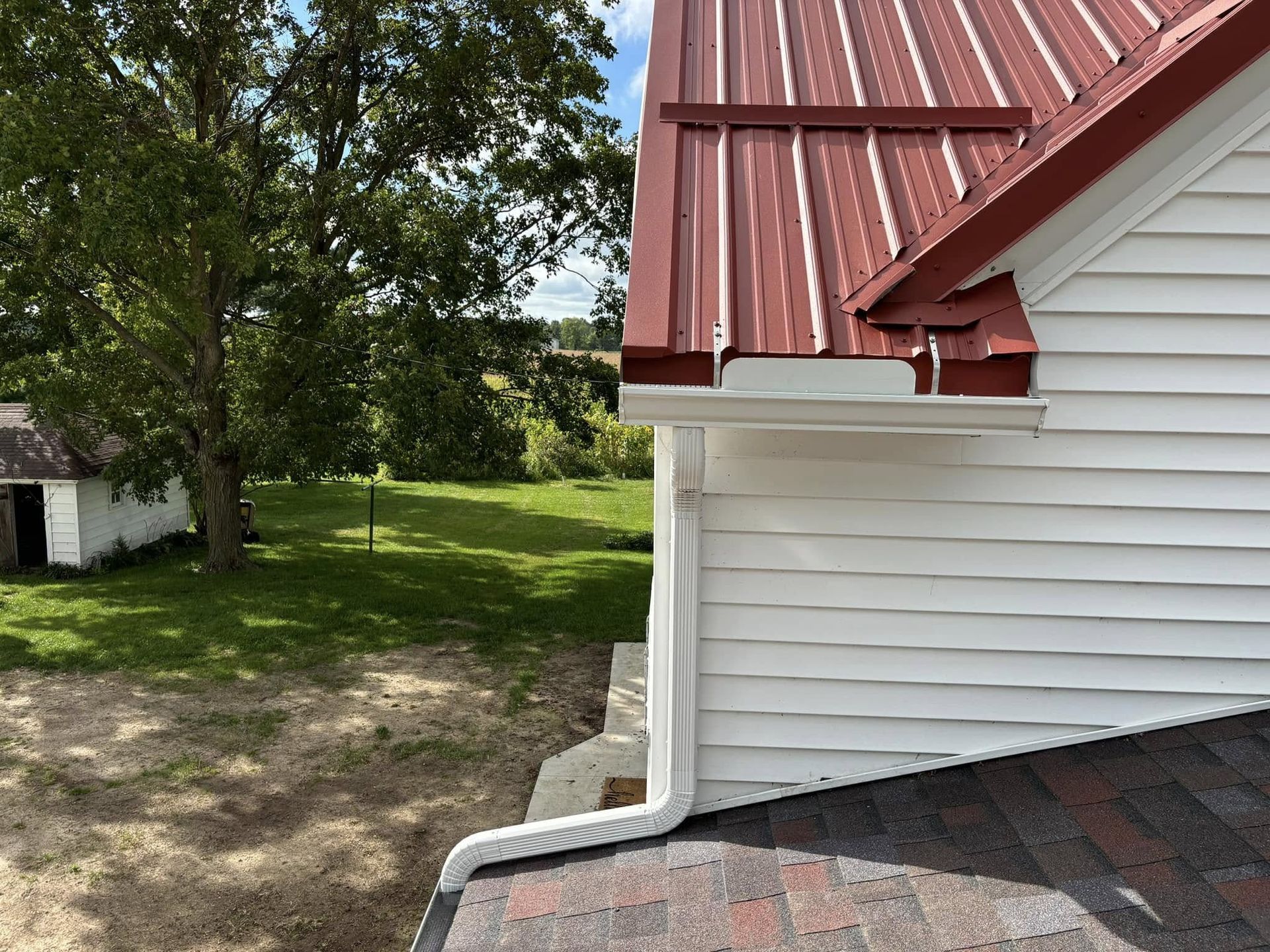 A view of a white house corner with a red metal roof, a white gutter system, and a downspout discharging onto a lower roof.