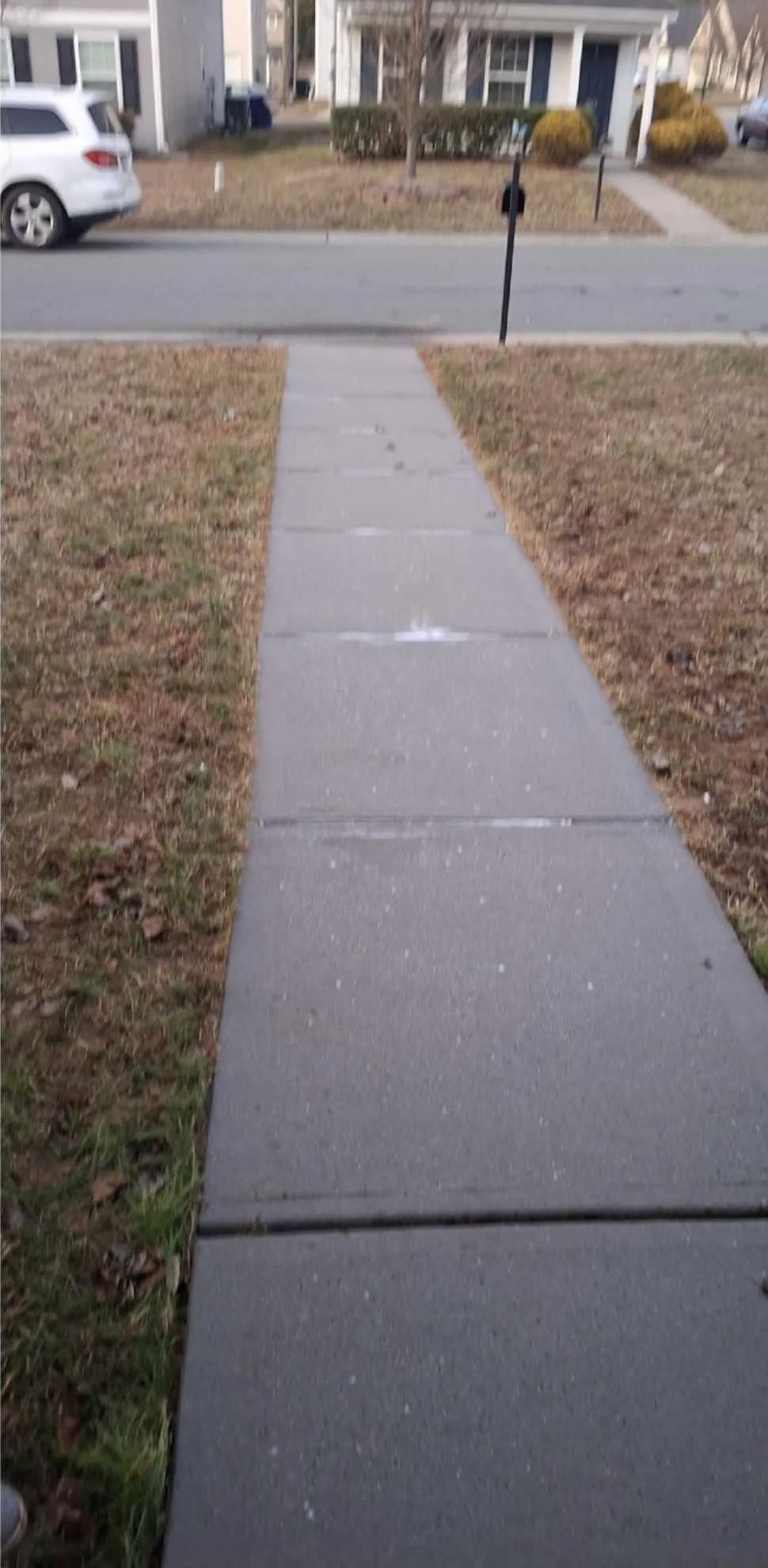 A concrete sidewalk leads toward a suburban street, flanked by brown grass and houses.