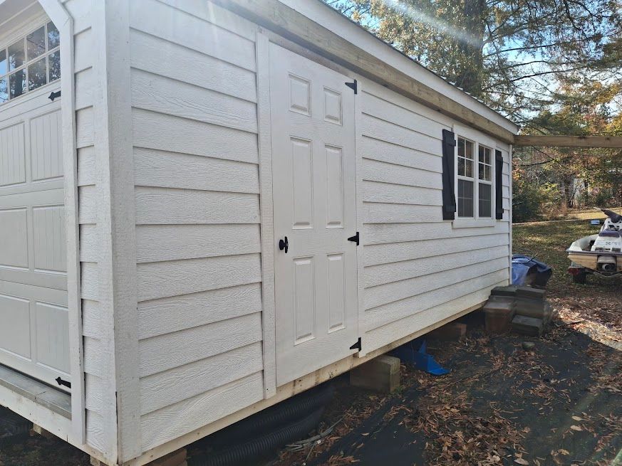 White shed with a door, window, and black shutters, on a wooden base in a yard.