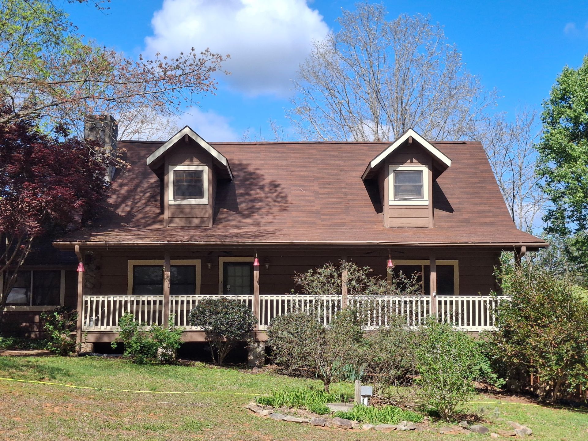 A rustic two-story wooden cabin with a wide front porch, white railings, and two dormer windows under a bright blue sky.