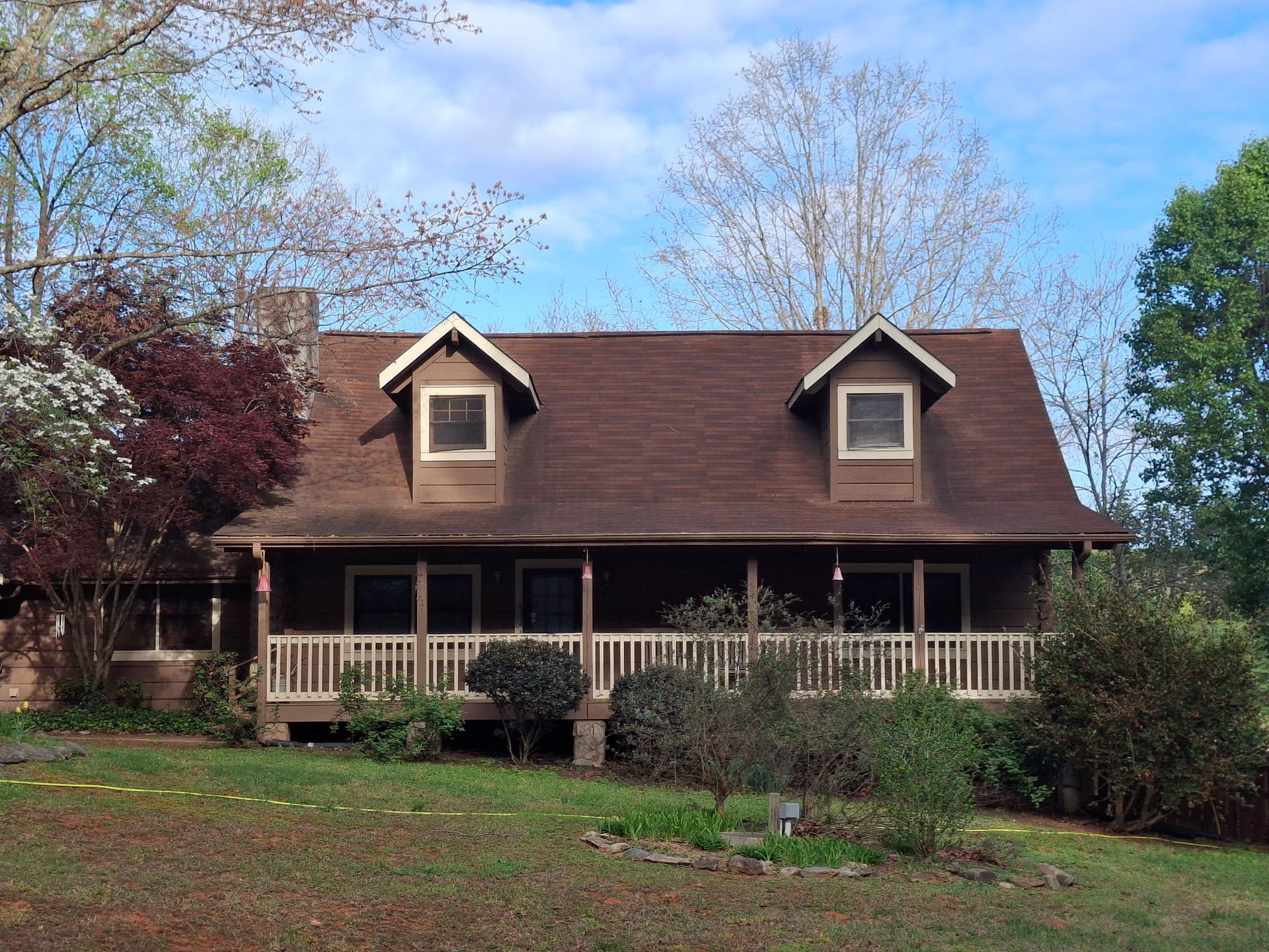 A brown, two-story wooden house with a front porch, two dormer windows, and a shingled roof, surrounded by trees.