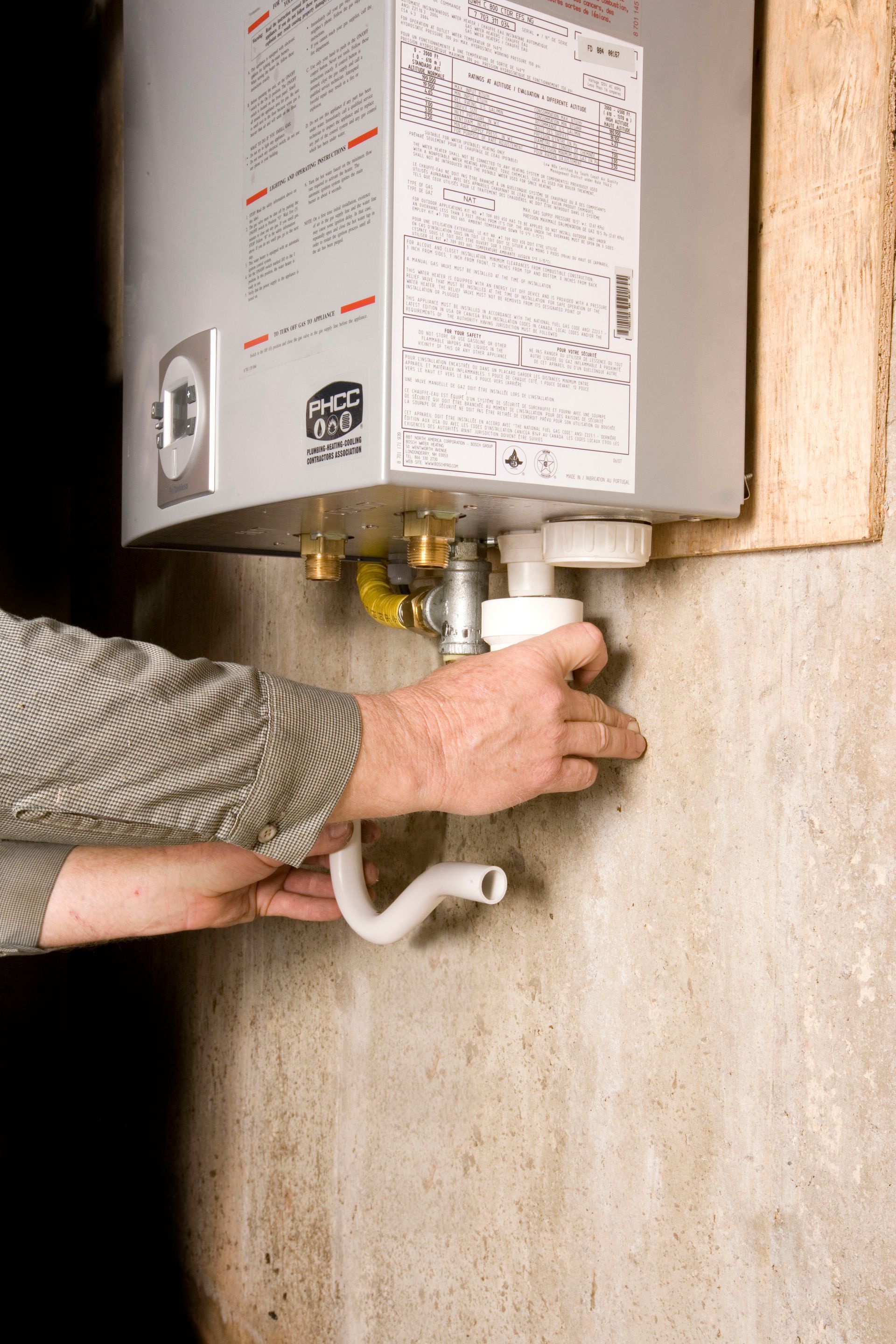 Person connecting a white hose to the bottom of a hot water heater against a concrete wall.