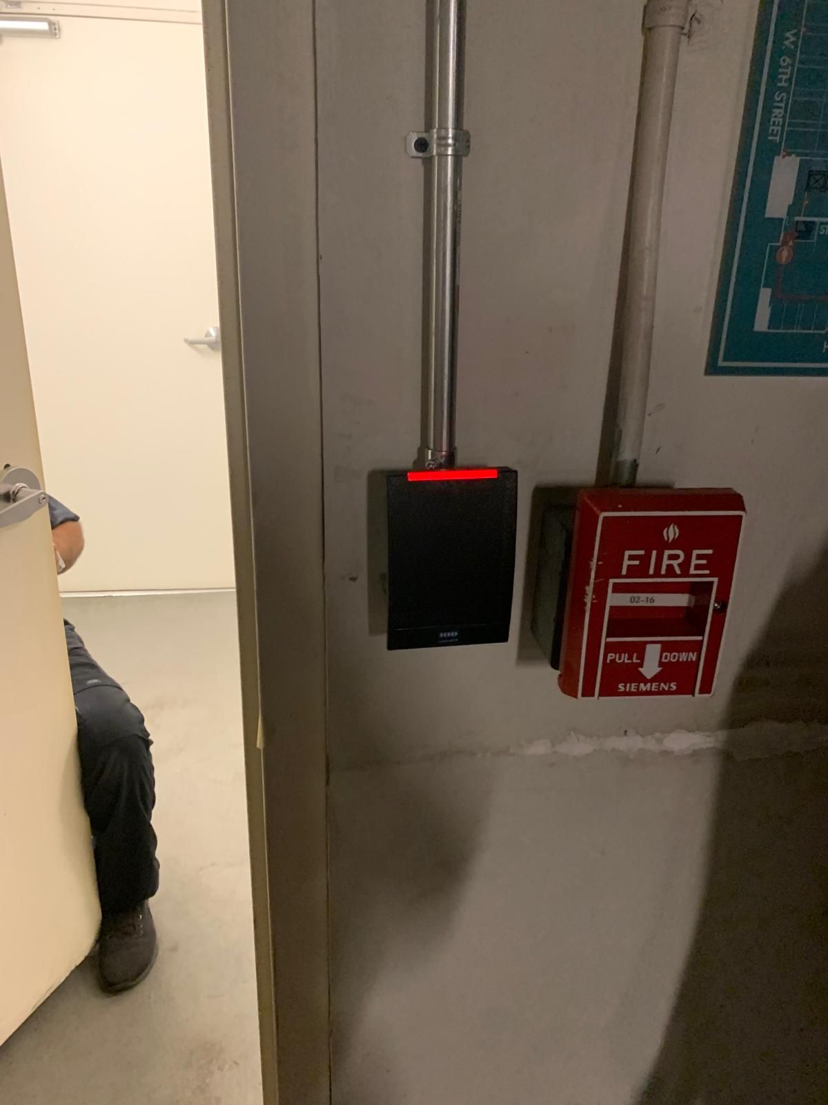 A black access card reader with a red light is mounted on a wall next to a red fire alarm pull station. A person is partially visible in the doorway.