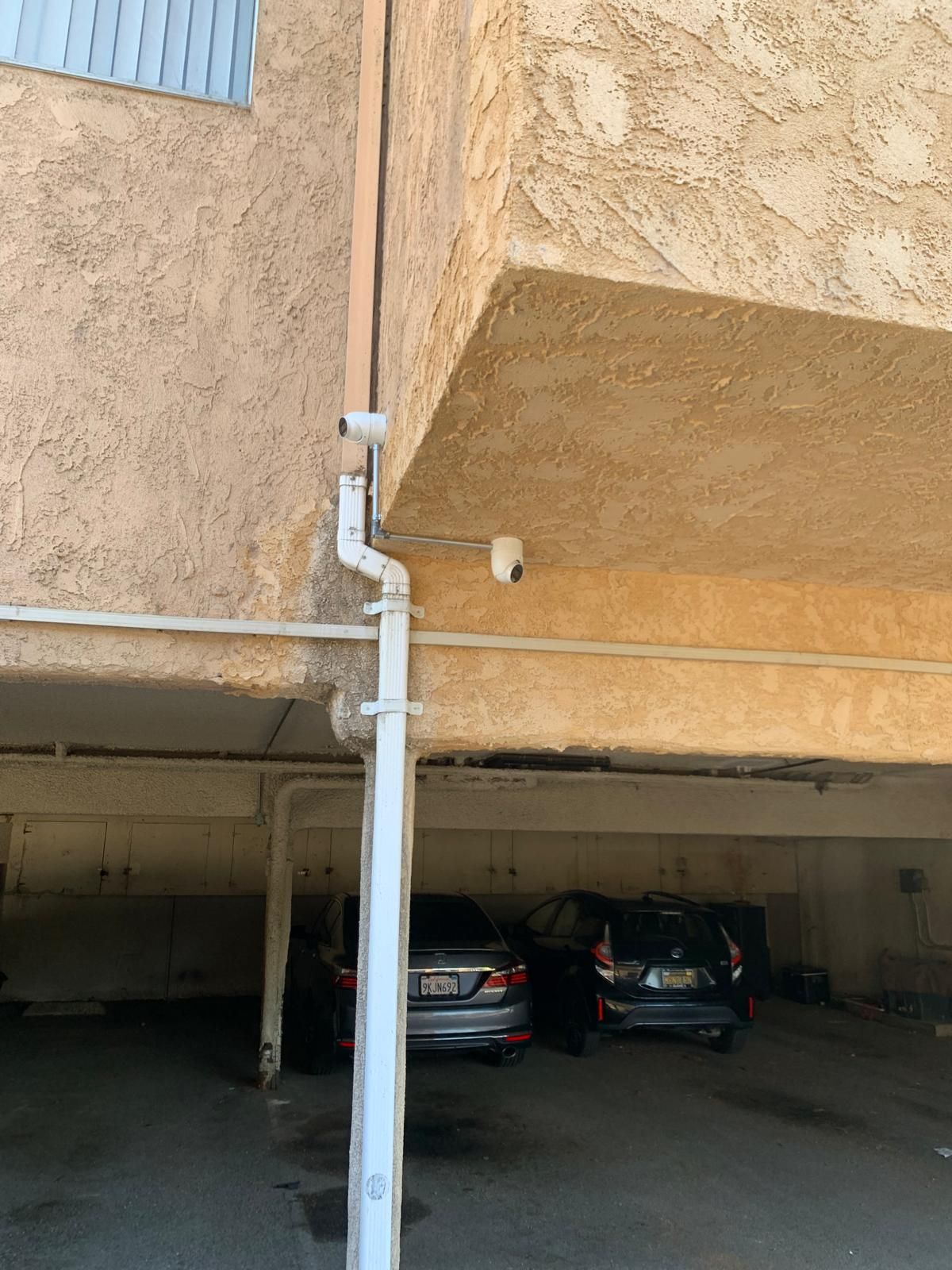 Security camera mounted on a building corner overlooking a parking garage with parked cars. A white rain gutter runs vertically.