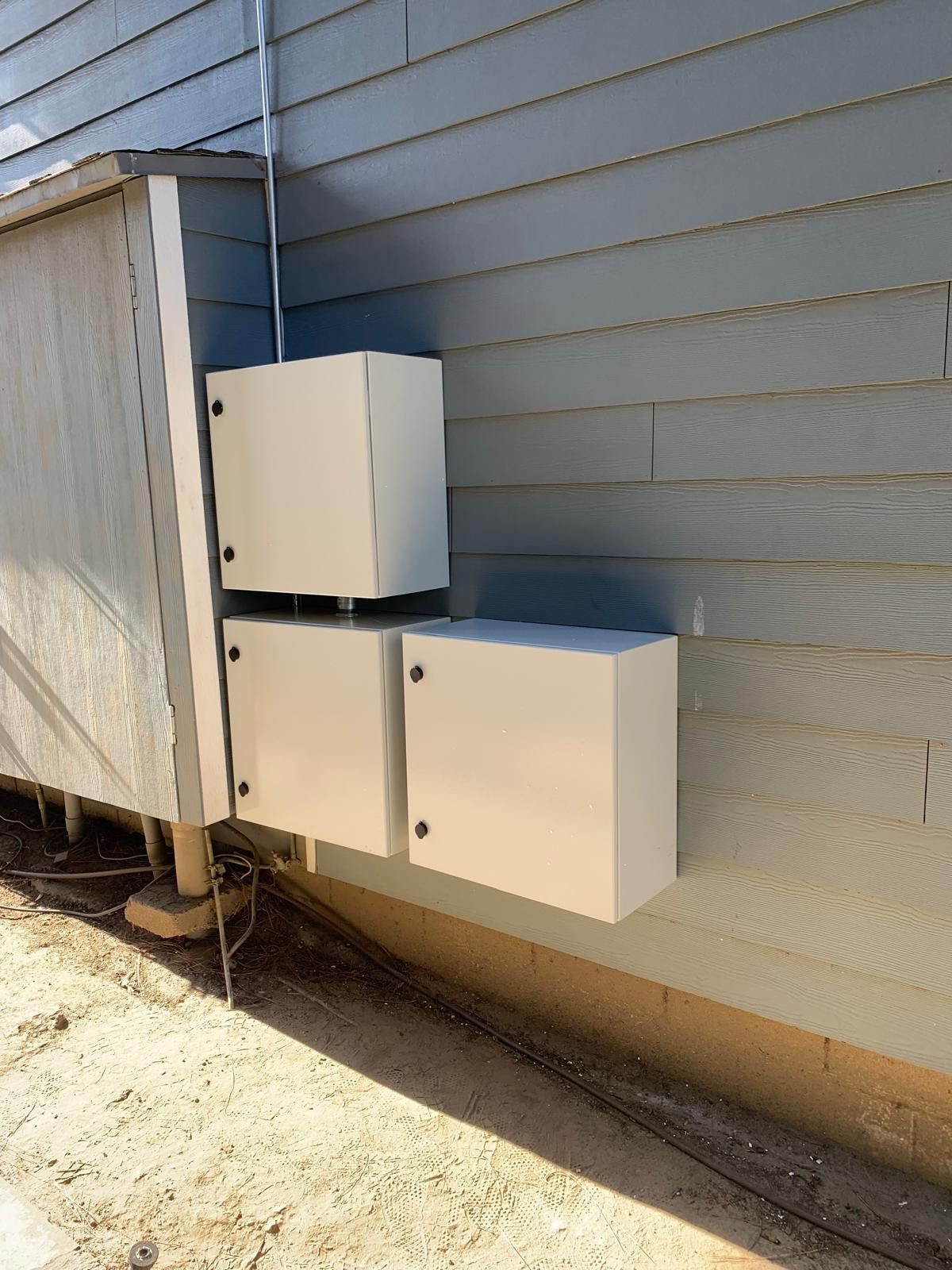 Three white electrical boxes mounted on a blue house exterior wall. A metal conduit runs upwards.