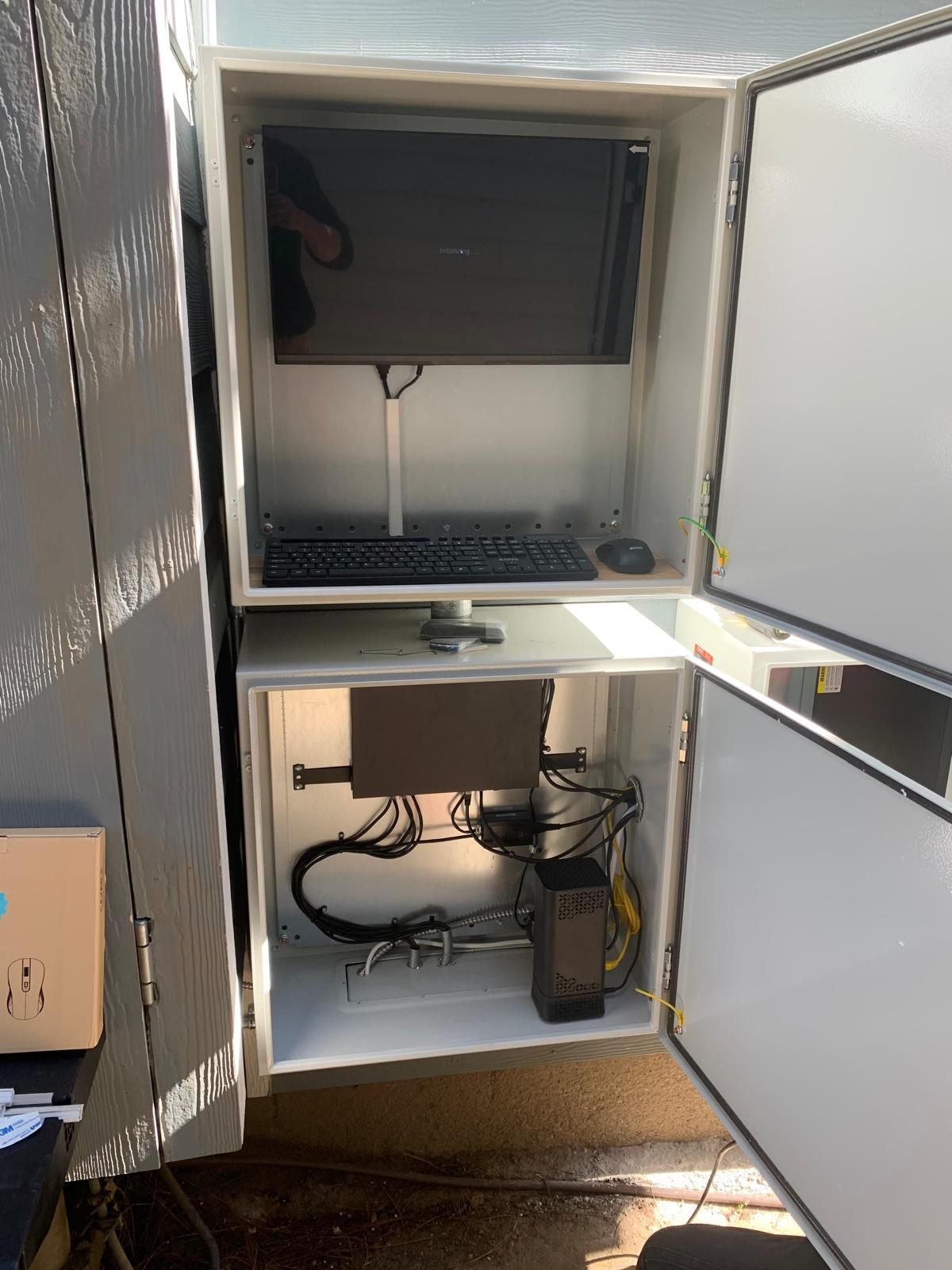 An outdoor enclosure with an open top containing a monitor, keyboard, and computer components; the lower section houses more electronics and wiring.