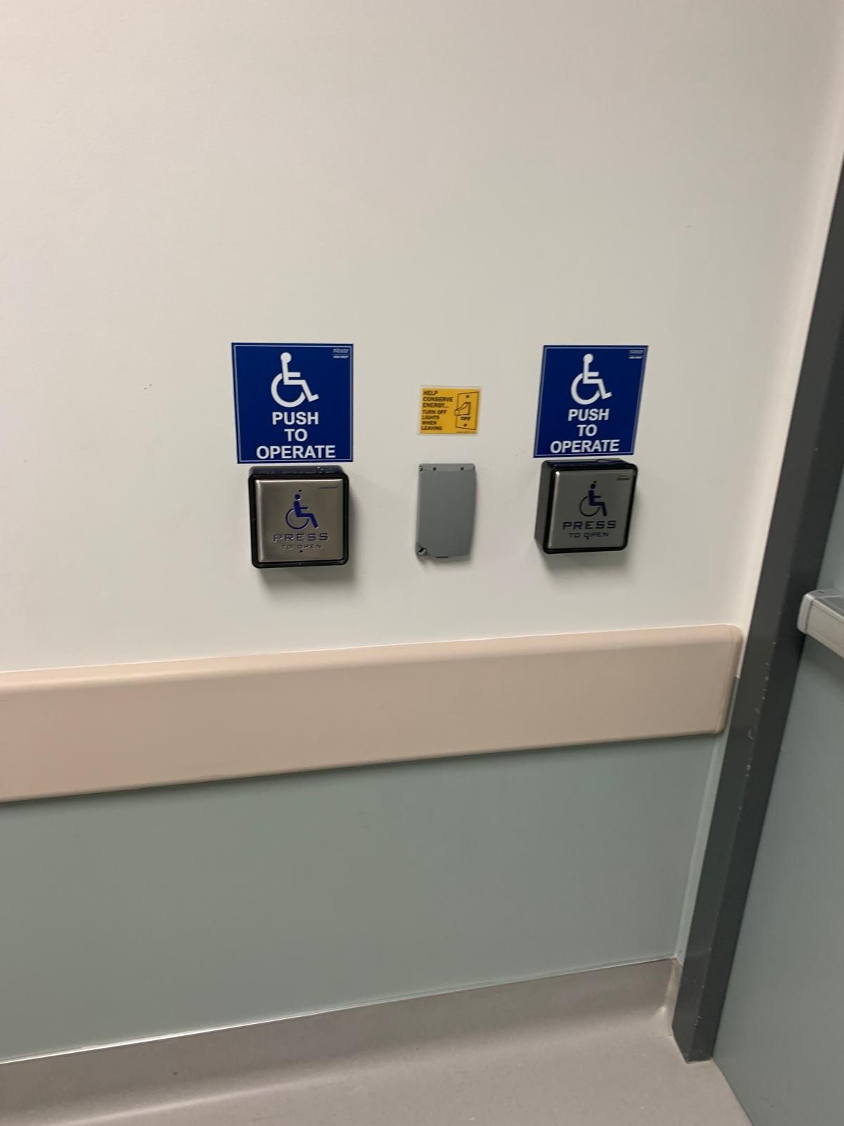 Wall with two blue accessible door opener buttons, centered between a neutral-colored switch and a yellow card. The buttons have the wheelchair accessible symbol and text: “PUSH TO OPEN”.