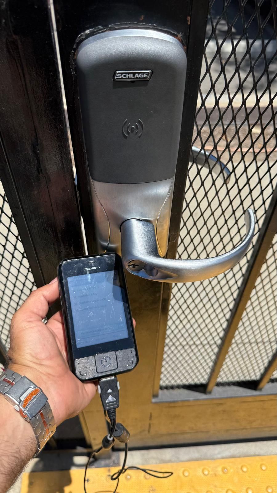 Person holding a phone near a gate lock. The lock is grey and silver. The gate is black and chain link.