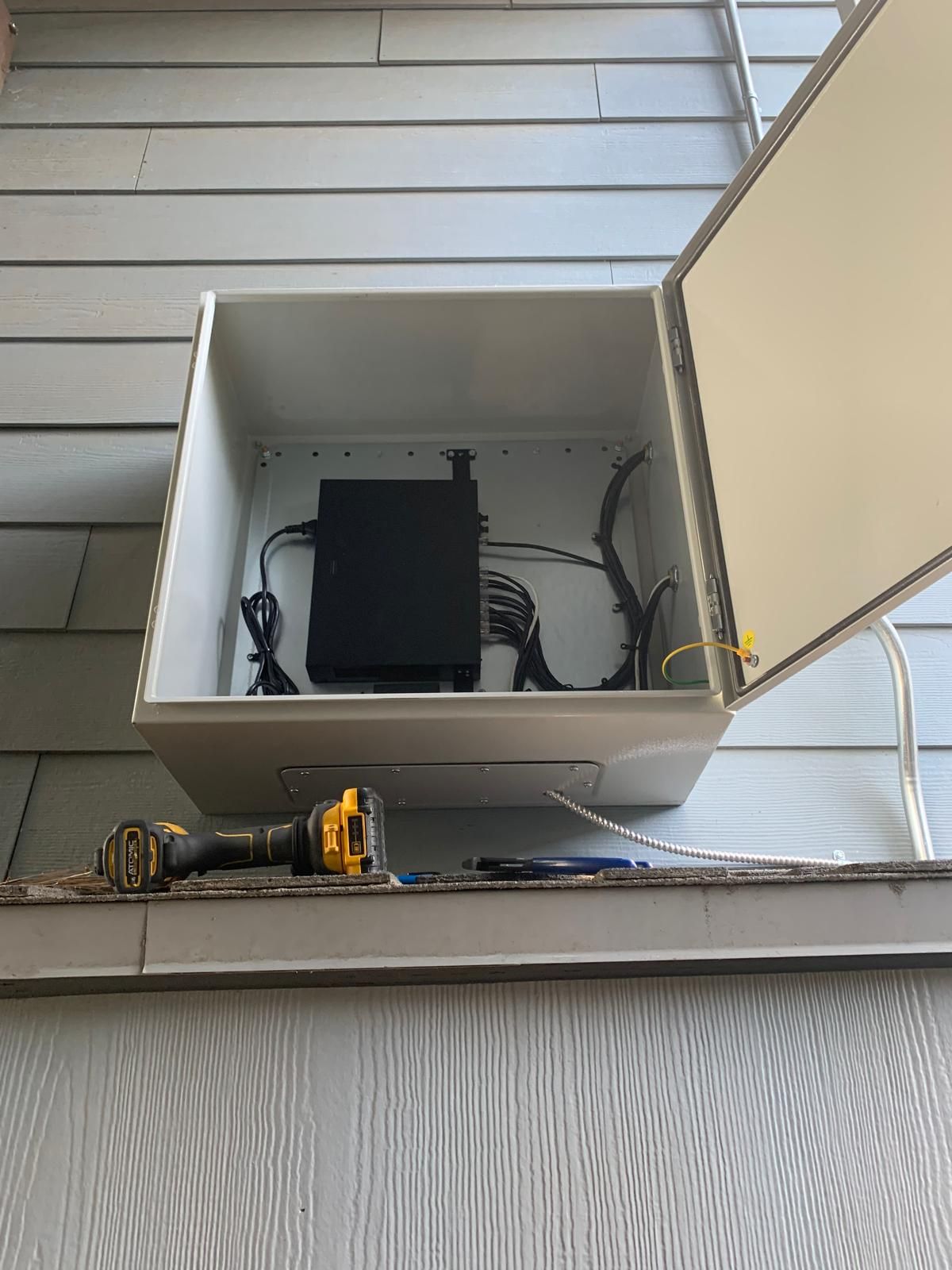 Open, white box on a light blue-gray building exterior. Inside, a black electronic device sits with wires. A drill rests on the ledge.