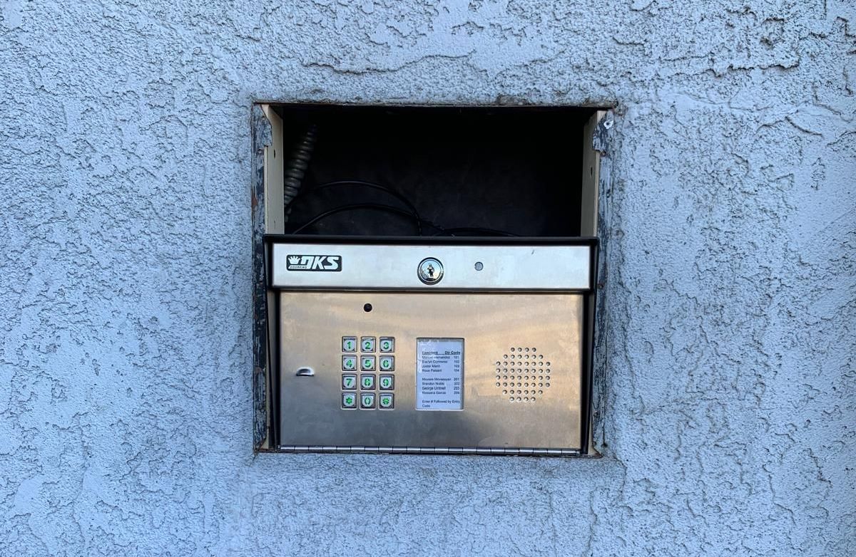 A silver entry system with a keypad and speaker is built into a stucco wall. The top has an open compartment.