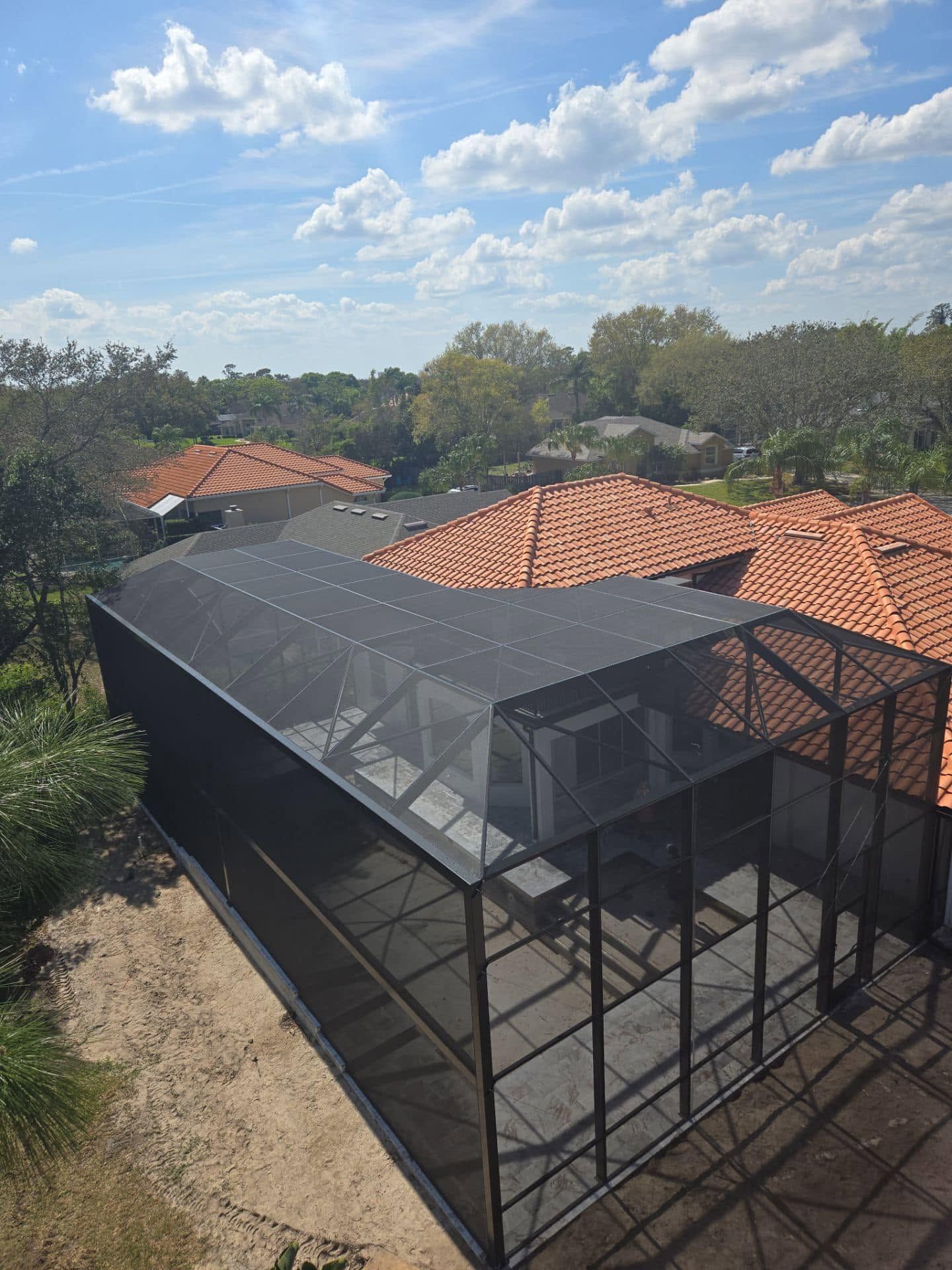 Black screened enclosure beside red-tile roofs under a bright blue sky with scattered clouds