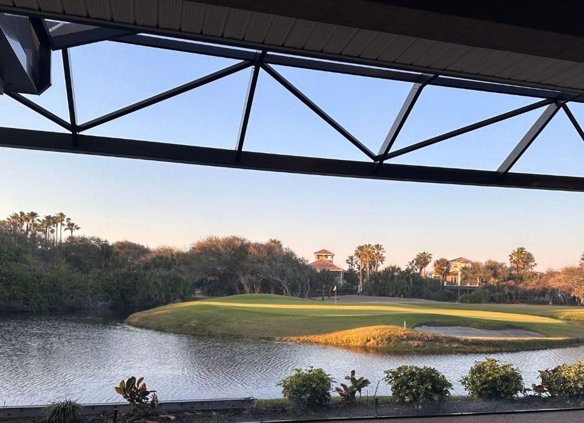 Sunlit golf green beside a pond, framed by a bridge and trees at sunset
