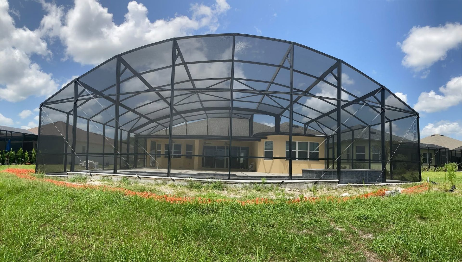 Large screened pool enclosure over a backyard, with grass and flowers in front under a cloudy sky