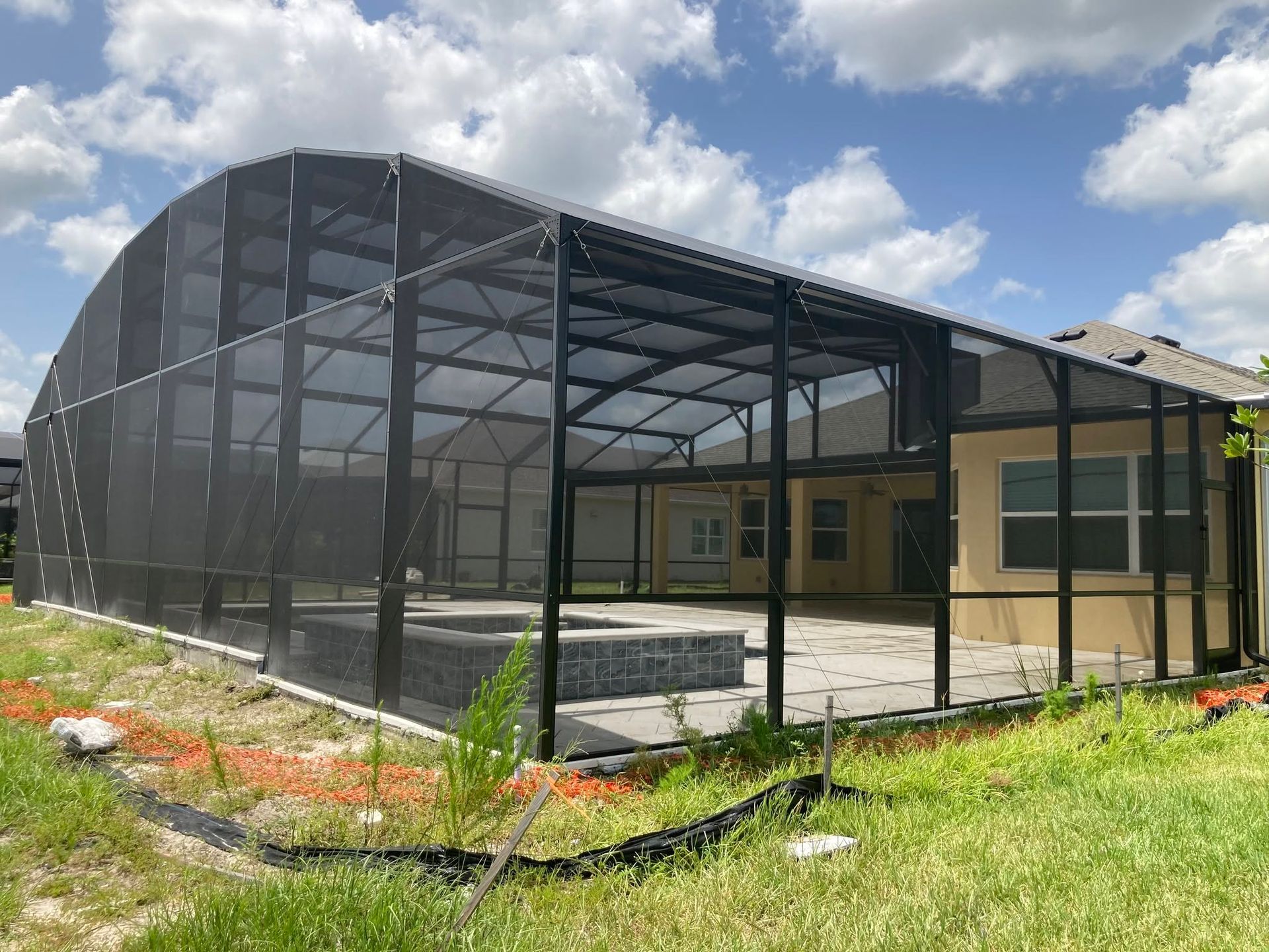 Mesh-enclosed patio or screened porch attached to a house, with black framing and grassy yard under cloudy sky