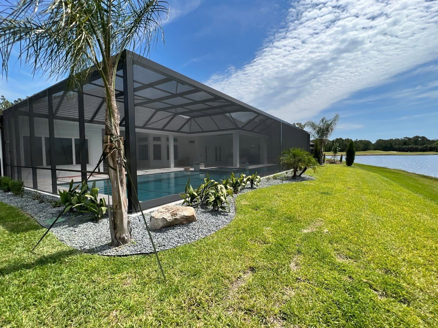 Screened backyard pool beside a lake, with green lawn, palm tree, and bright blue sky