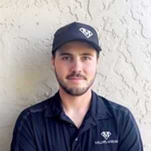 Man in a dark cap and shirt stands against a textured wall, facing the camera.