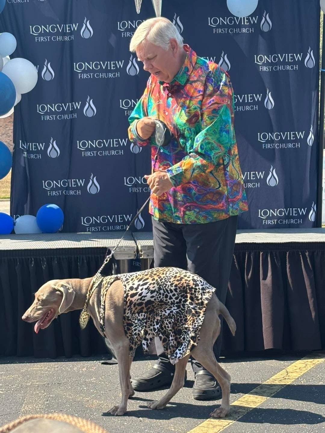 A man is standing next to a dog wearing a leopard print dress.