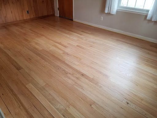 Light-colored hardwood floor in a room with a window and wooden paneling on one wall.