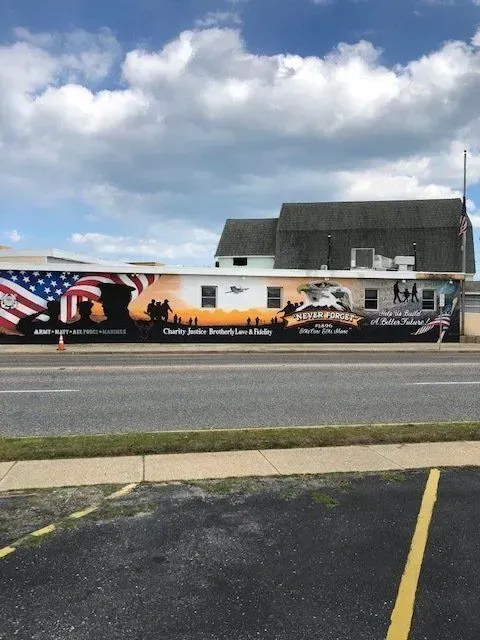 Mural on a building: American flag, soldiers, tanks, and text honoring veterans. Overcast sky.