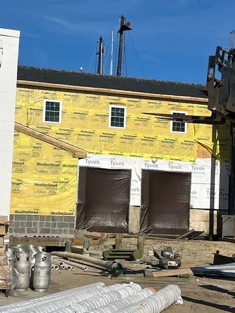 Building under construction, yellow wrap, windows, large garage doors. Construction equipment and materials in the foreground, blue sky.