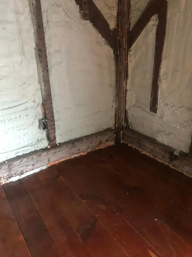 Interior corner of a room, showing exposed dark brown wooden beams, light tan walls, and a wood floor.