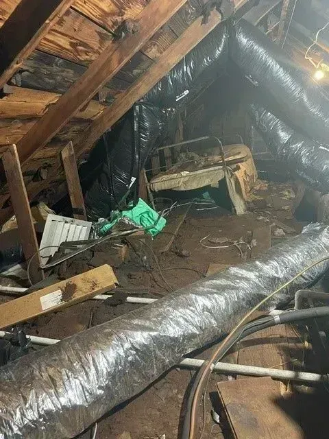 Attic filled with debris, insulation, and HVAC ductwork, suggesting disrepair and clutter.
