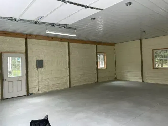 Interior of a garage with spray foam insulation on the walls. Gray concrete floor, white ceiling, and wooden beams are visible.