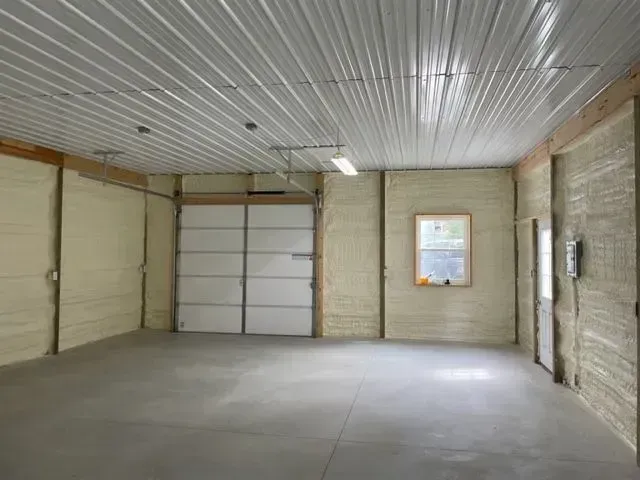 Interior view of a garage with spray foam insulation on walls, white garage door, and a window.
