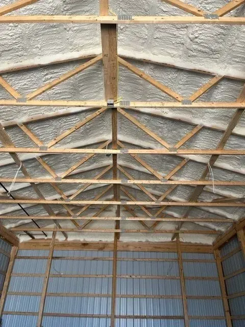 Interior of a building with wooden trusses and spray foam insulation. Metal siding is visible.