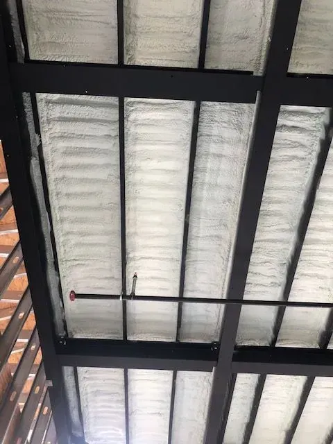 Metal roof structure with spray foam insulation; black beams, a sprinkler pipe, and a brick wall visible.