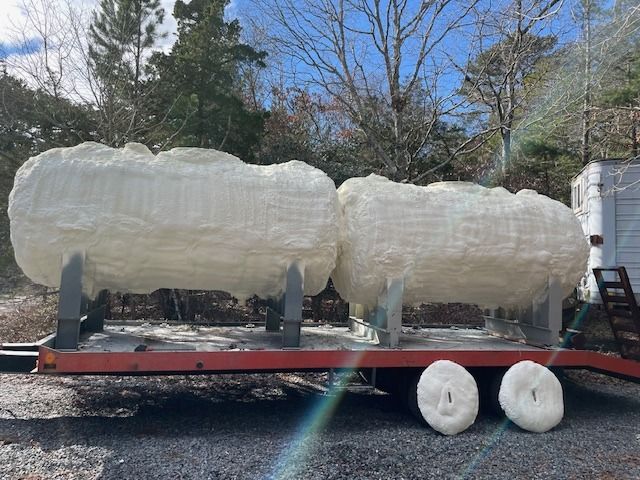 Two large tanks, covered in white insulation foam, on a flatbed trailer. Two foam cut-offs rest on the ground.