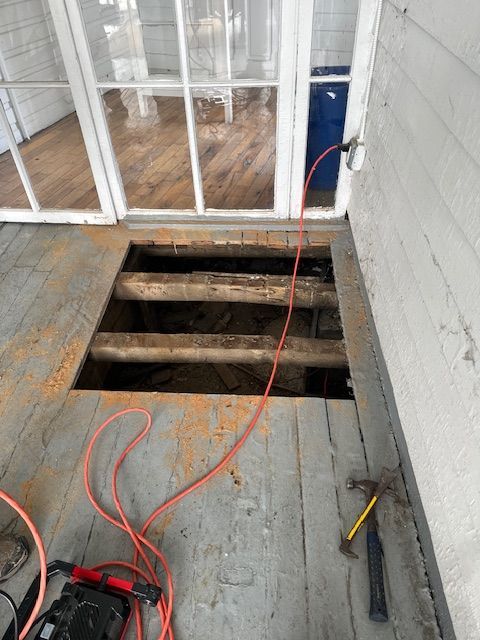 Hole in porch floor with exposed support beams. Orange extension cord and tools are present. White trim and windows in the background.