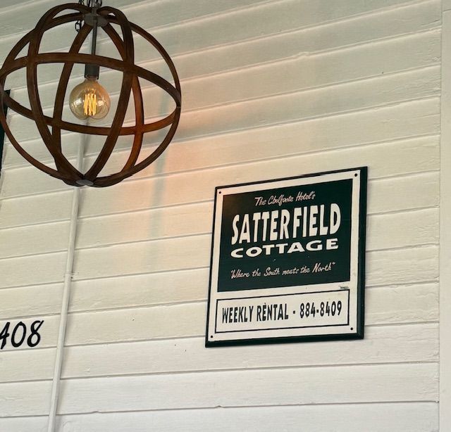 Sign for Satterfield Cottage, weekly rental, on white siding, under orb light fixture.