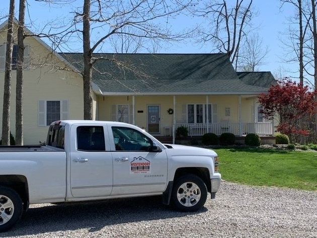 A white service pickup truck parked on a gravel driveway in front of a yellow house with a covered porch.