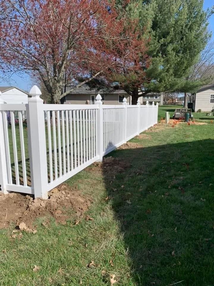 A white vinyl picket fence stands in a grassy backyard with disturbed earth along its base and trees in the background.